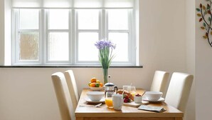 Une table de petit-déjeuner avec un bol de fruits, un vase de fleurs et une cafetière.