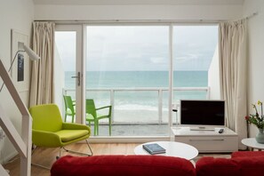 Une pièce dotée d’une grande fenêtre donnant sur la mer, d’un fauteuil vert, d’un canapé rouge, d’un meuble TV blanc et d’une petite table sur laquelle se trouve un livre.