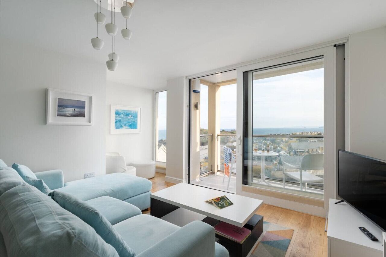 Sala de estar moderna com sofá, mesa de centro e televisão. Há uma varanda com vista para a cidade.