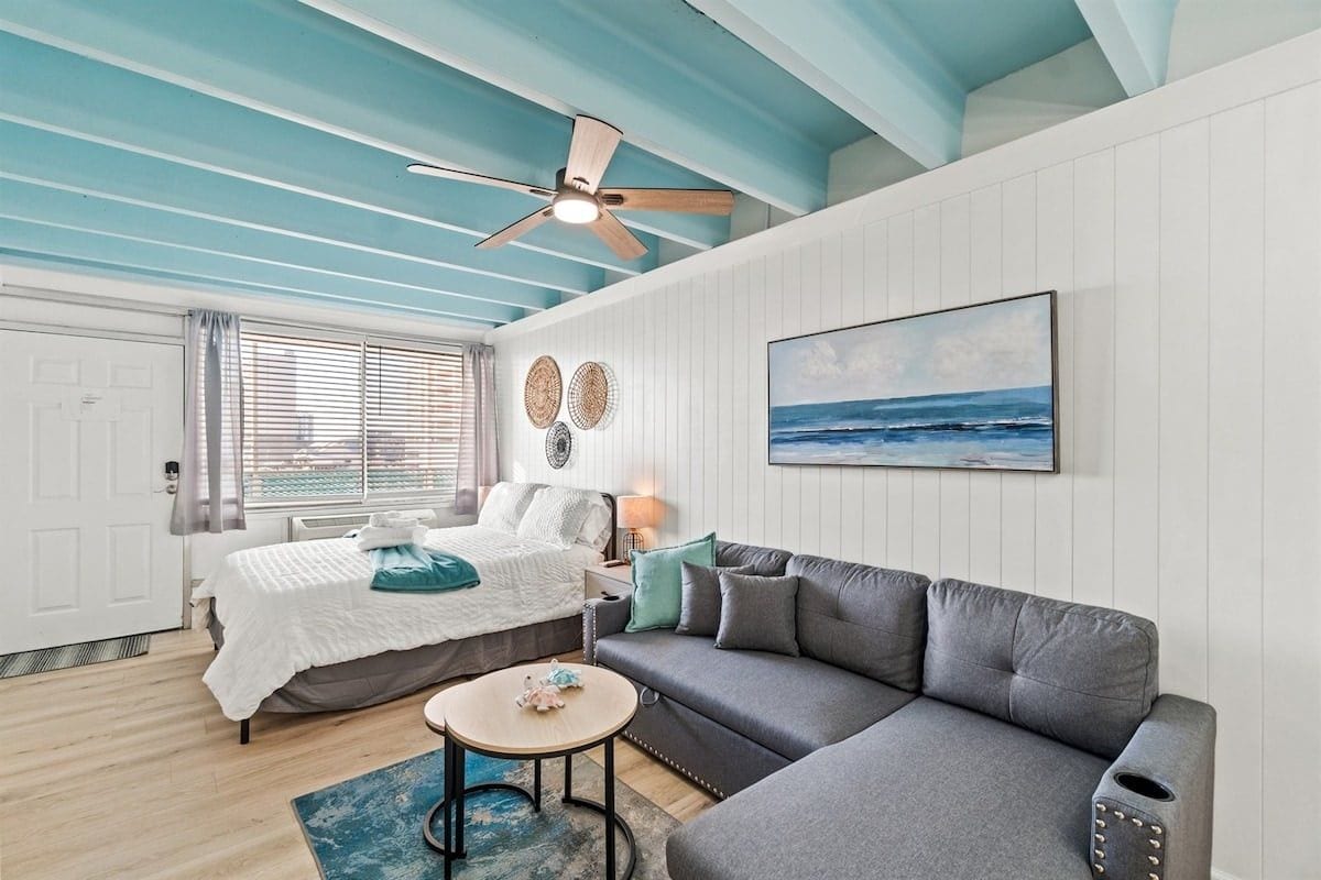 A room with a bed, a gray sofa, a round coffee table, a ceiling fan, and a painting of the ocean.