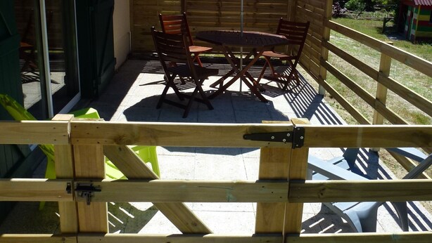 Una recinzione in legno con tavolo e sedie sotto un ombrellone, un pontile di legno e sullo sfondo alberi e un edificio.