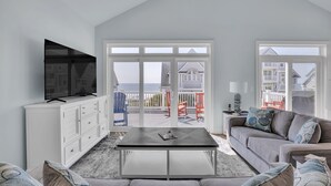 Een moderne woonkamer met een grote televisie, een bank en een salontafel. Er is een balkon met uitzicht op zee en een huis op de achtergrond.