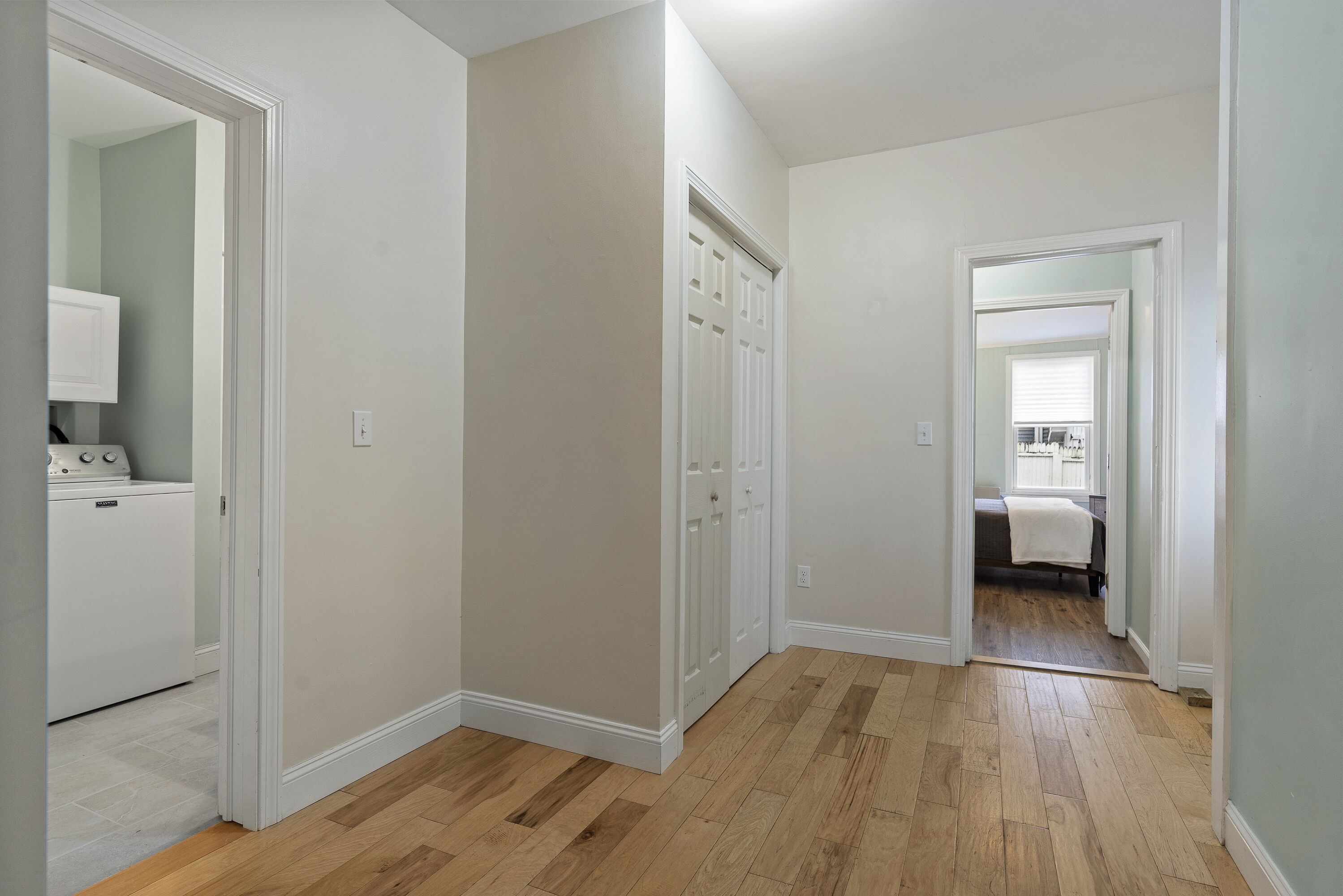 A hallway with wooden flooring, white walls, and a door leading to a bedroom with a bed and window.