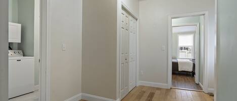 A hallway with wooden flooring, white walls, and a door leading to a bedroom with a bed and window.