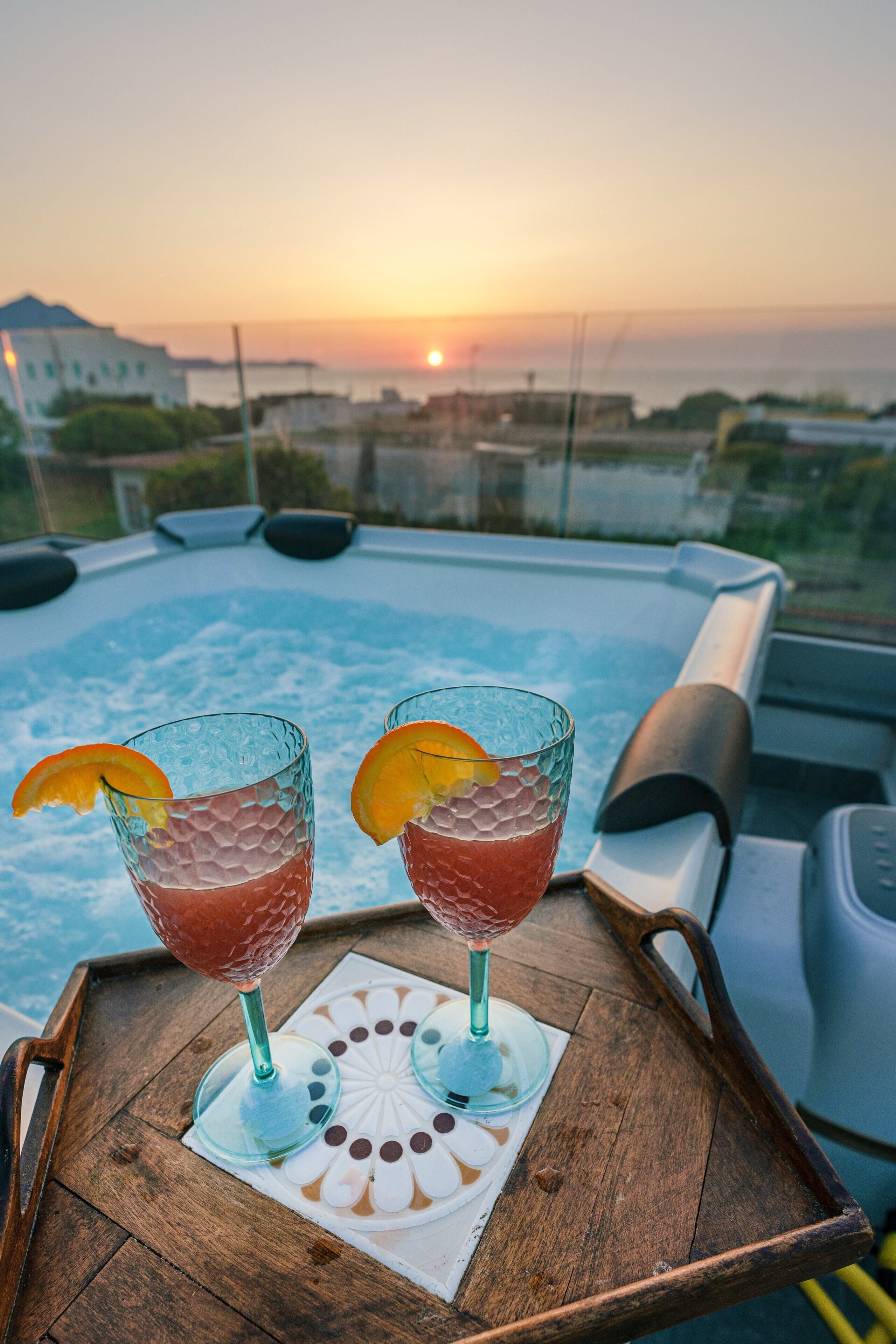 Un jacuzzi con dos vasos de bebida roja sobre una bandeja de madera, con vista a un atardecer.