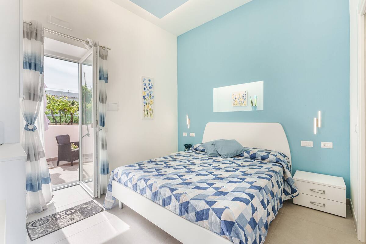 A bedroom with a bed, a nightstand, a window with curtains, and a view of the outdoors.