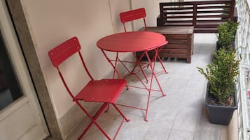 Un balcón con sillas metálicas rojas, un banco de madera, plantas en macetas y piso de baldosas.