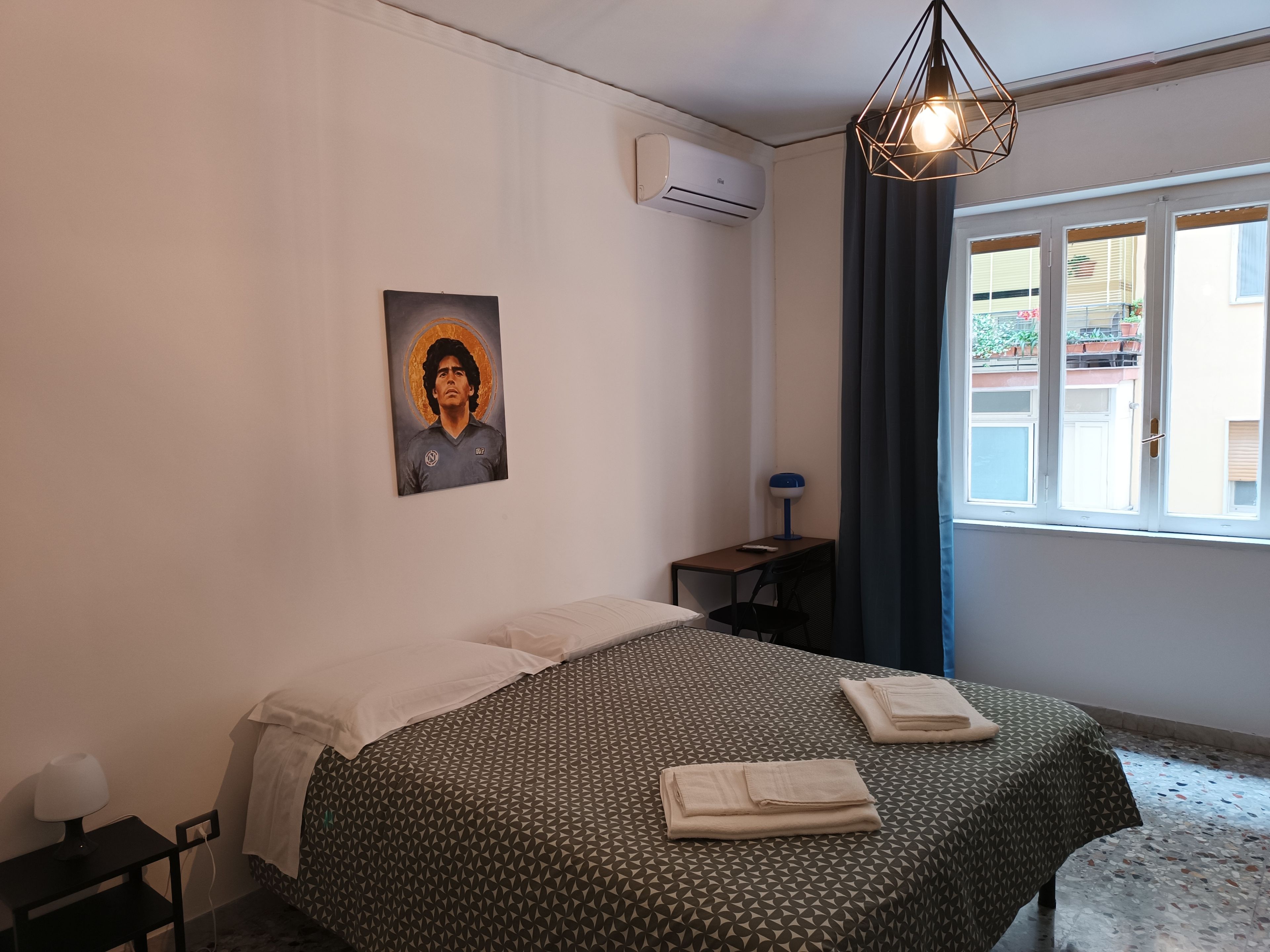 Quarto de hotel com cama, escrivaninha, cadeira, luminária, pintura, ar condicionado e janela com cortinas.
