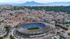 Et stadion med stor tilskuerkapacitet, omgivet af et tæt bebygget byområde med forskellige bygninger og et bjergrigt baggrundslandskab.