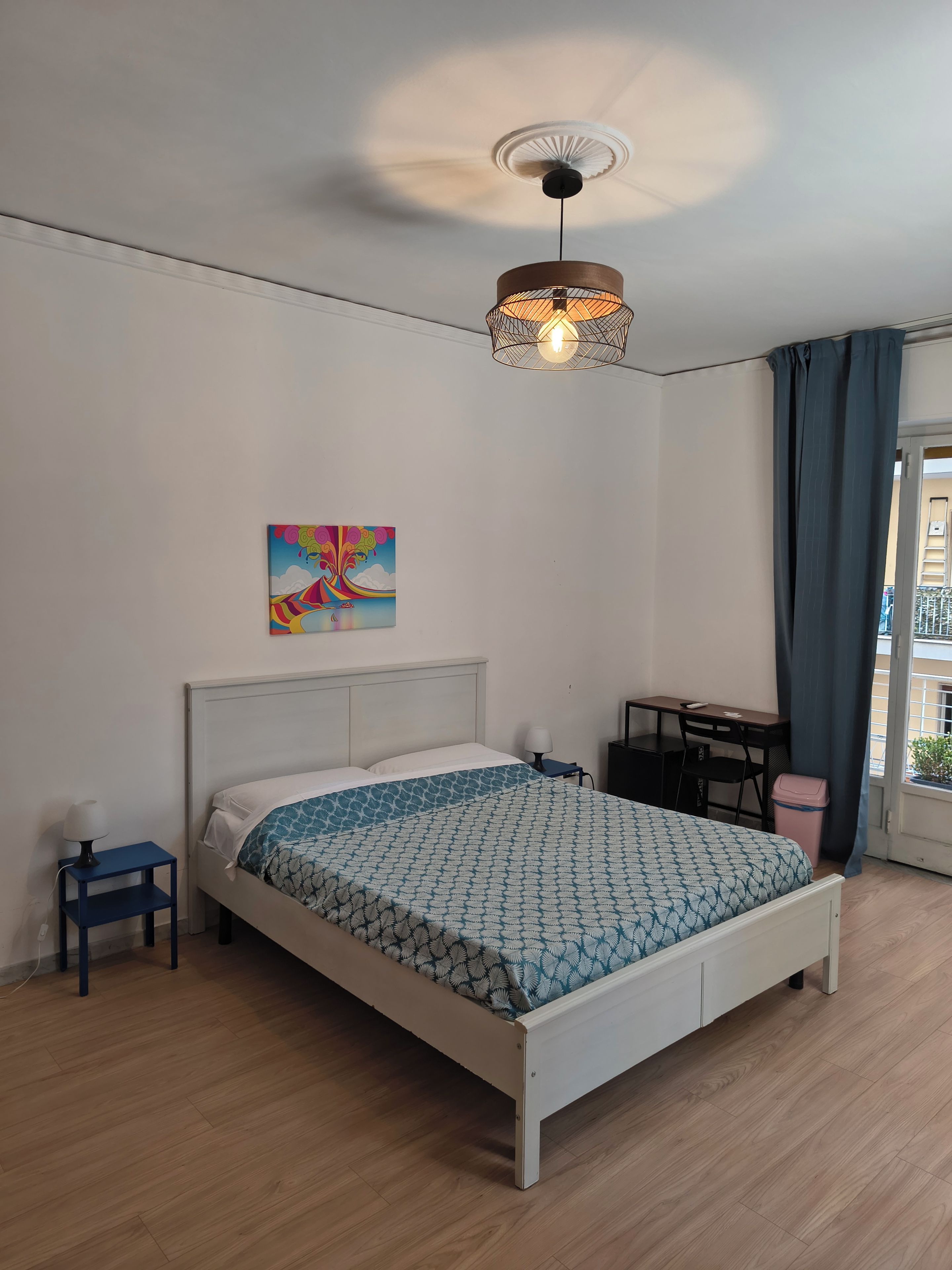 Un dormitorio con cama, escritorio, silla, lámpara, ventilador de techo y ventana con cortinas.