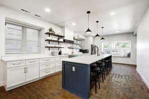 Una cocina moderna con armarios blancos, una isla central, iluminación colgante y suelo de ladrillo.