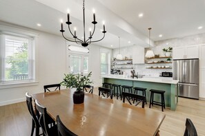 A modern kitchen with a dining area, featuring a chandelier, a central island with stools, and a stainless steel refrigerator.