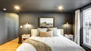 Ein Schlafzimmer mit einem großen Bett, zwei Nachttischlampen, einem gerahmten Bild an der Wand und einem Fenster mit Vorhängen.