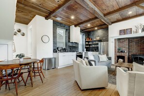 A modern kitchen with white cabinets, a dining area with a wooden table and chairs, and a living room with a white sofa and fireplace. - The Nook at Legacy Manor (Lancaster)