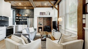 Una sala de estar moderna con chimenea, vigas de madera en el techo y una zona de cocina con salpicadero de piedra.