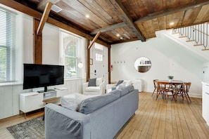 Un salon moderne avec un canapé gris, des fauteuils blancs, un téléviseur et un coin repas comprenant des chaises et une table en bois.