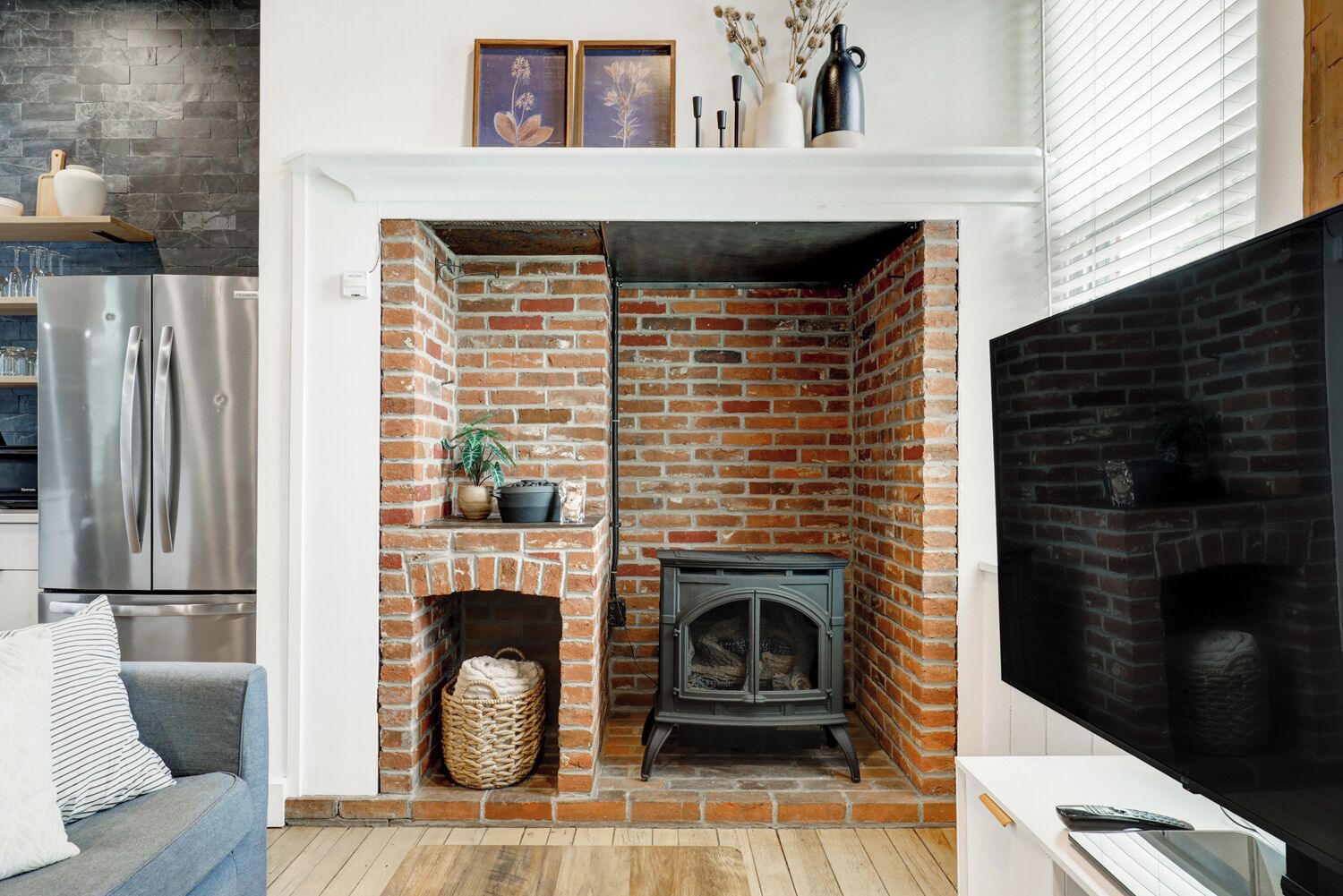 A living room with a brick fireplace, a television, and a modern kitchen with a stainless steel refrigerator.
