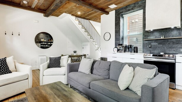 Una sala moderna con un sofá gris, sillones blancos y una mesa de centro de madera. Se ve una cocina con estufa y armarios al fondo.