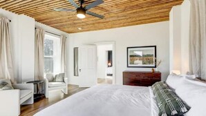 Un dormitorio con una cama grande, un ventilador de techo, techo de madera, un aparador y una ventana con cortinas.