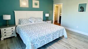 Une chambre à coucher comprenant un lit, deux tables de chevet, un ventilateur de plafond et une porte donnant sur une autre pièce.