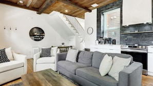 Un salon moderne avec un canapé gris, des fauteuils blancs et une table basse en bois. On aperçoit une cuisine avec une cuisinière et des placards à l’arrière-plan.
