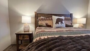 Une chambre avec un grand lit, deux tables de chevet avec des lampes et une couvre-lit à motifs forestiers.