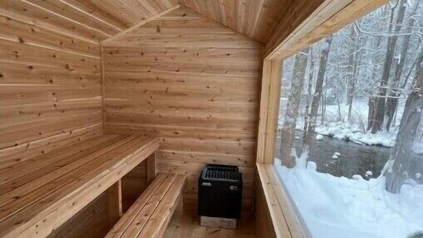Un sauna en bois, avec des bancs et un poêle noir, et une fenêtre donnant sur une forêt enneigée.