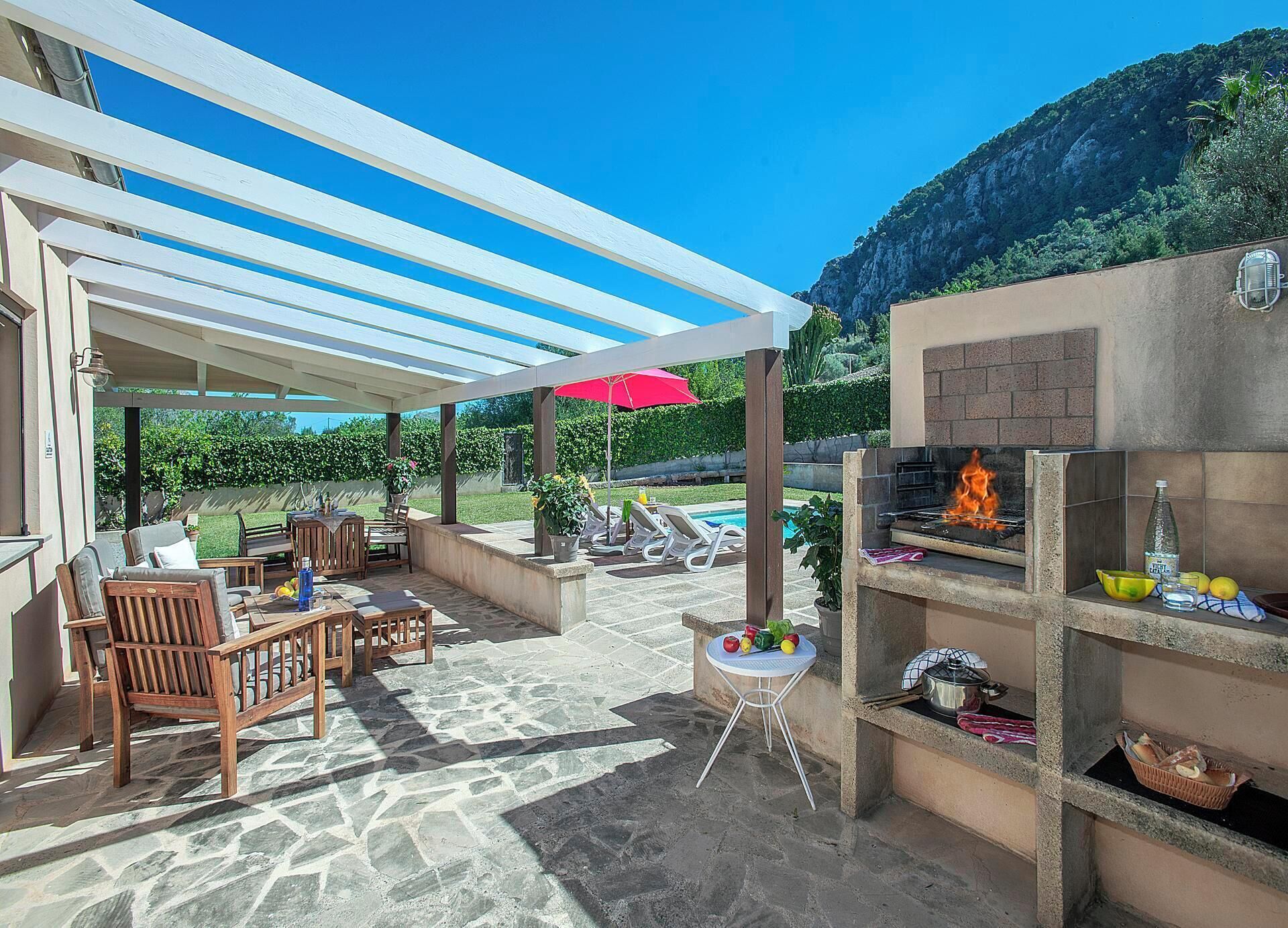Eine Terrasse im Freien mit Steinboden, Holzmöbeln, einem Grill und einem Pool im Hintergrund.