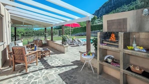 An outdoor patio with stone flooring, wooden furniture, a grill, and a pool in the background. - Ca Na Lida - Two Bedroom Villa, Sleeps 4 (Mallorca)
