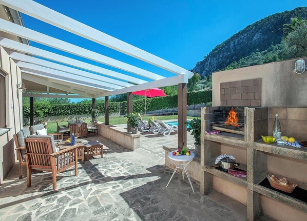 An outdoor patio with stone flooring, wooden furniture, a grill, and a pool in the background. - Ca Na Lida - Two Bedroom Villa, Sleeps 4 (Mallorca)