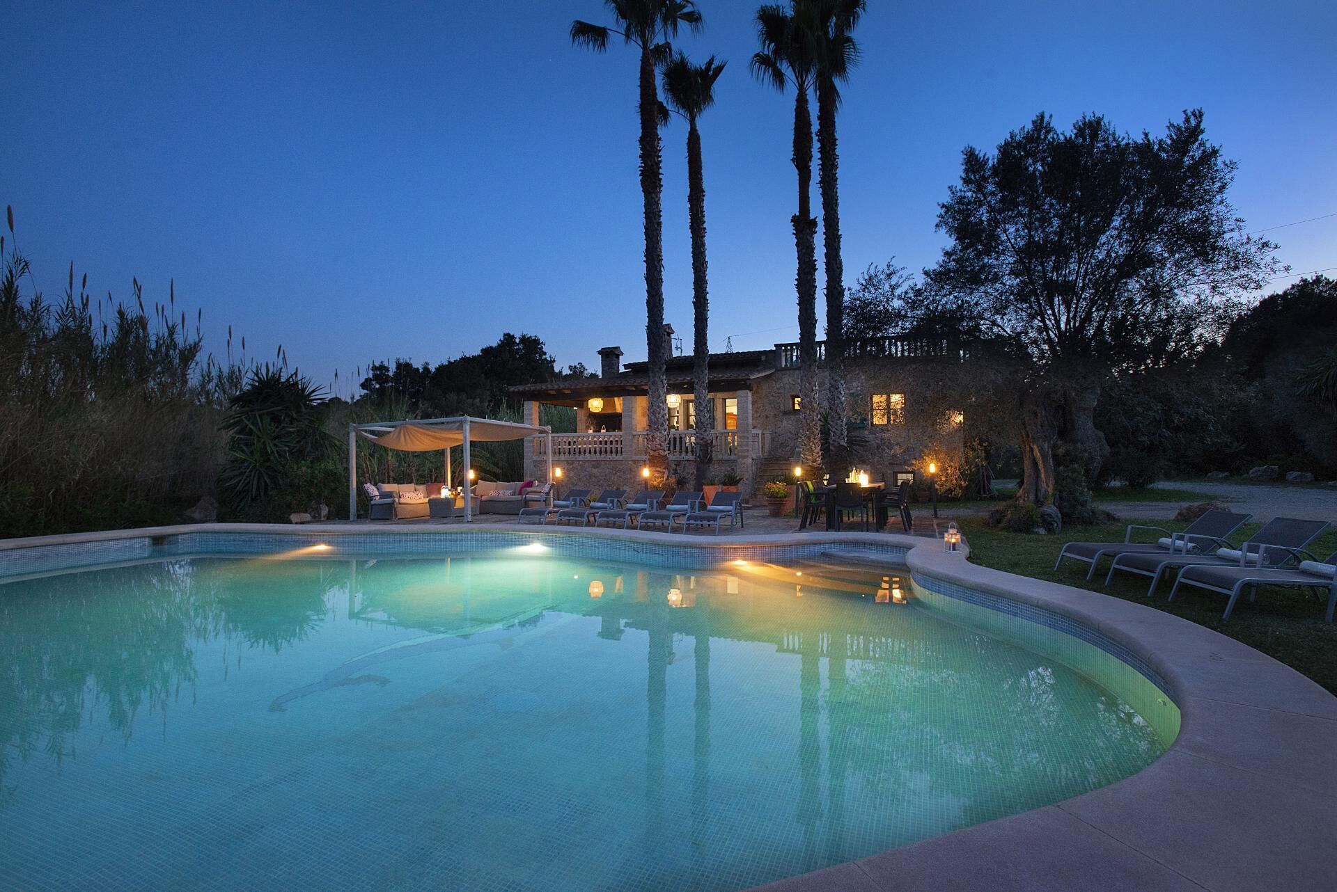 A swimming pool with lounge chairs and a covered seating area, surrounded by tall palm trees and a stone building with outdoor lighting.
