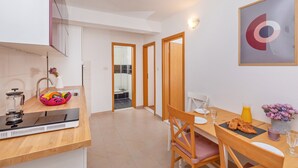 Una cocina moderna con estufa integrada, un frutero y un lavamanos. Hay una zona de comedor con una mesa preparada para dos personas, un jarrón de flores y un vaso de jugo de naranja.
