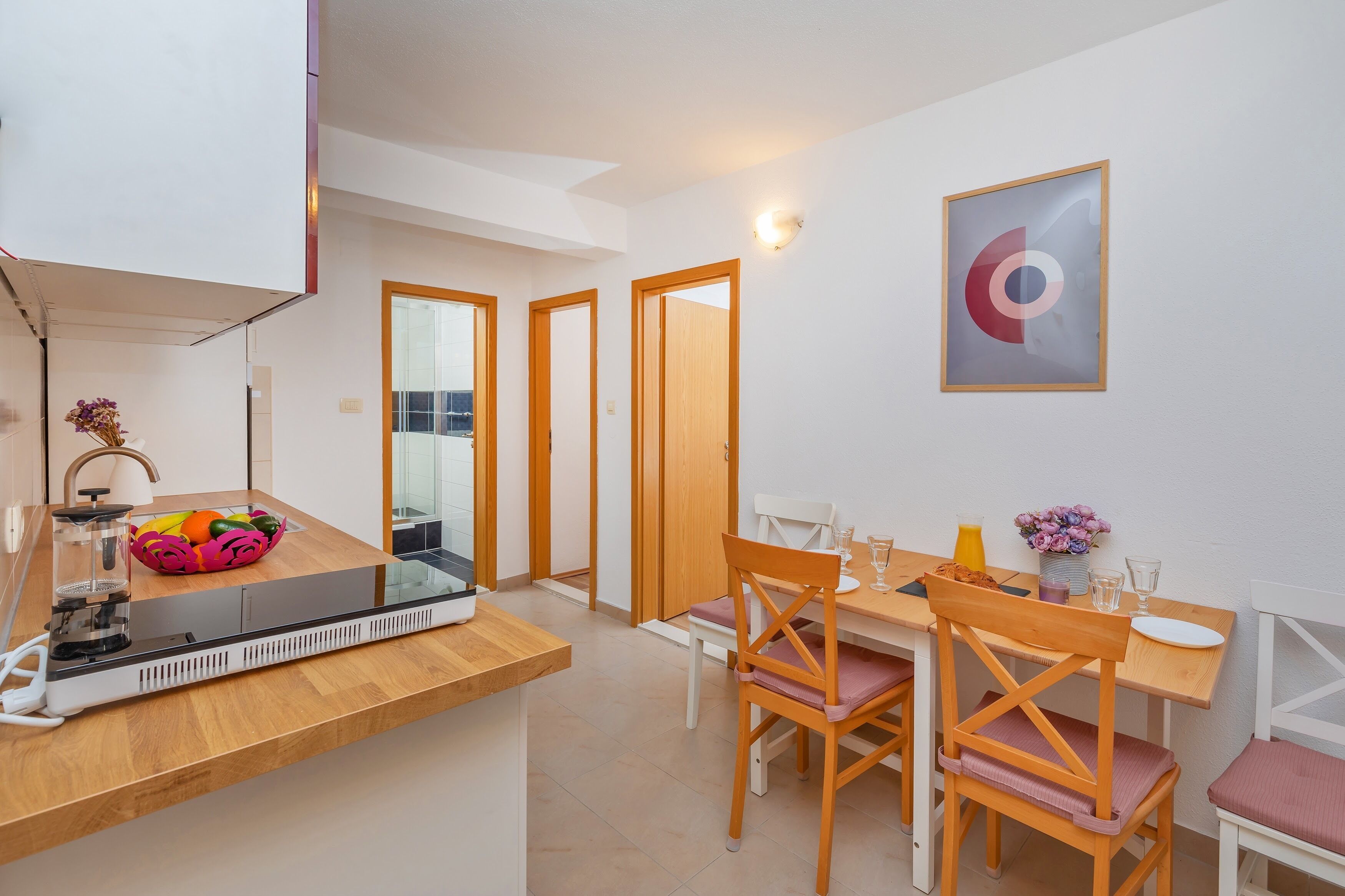 A modern kitchen with a wooden countertop, a sink, and a fruit bowl. There is a dining area with a table, chairs, and a vase of flowers.