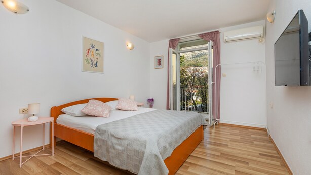 Un dormitorio con piso de madera, una cama con manta gris, una mesita de noche con lámpara y una ventana con vista a árboles.