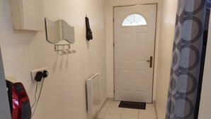 Un couloir avec une porte blanche, un radiateur, une prise de courant et un miroir au mur.