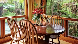 A wooden dining table with chairs, a hanging light fixture, and a view of a lush green forest through large windows. - The Pacifica Inn/ The Gecko and Volcano Room (Volcano)