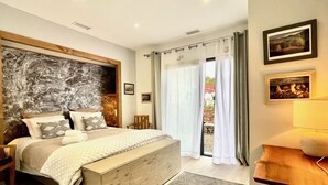 A bedroom with a large bed, a wooden headboard, a bedside table, a lamp, and a view through the window. - Les Terres Rouges du Salagou 5min du lac ! Natura 2000 , Atelier D'artiste (Le Bosc)