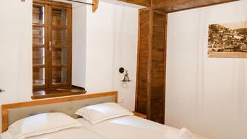 Un dormitorio con dos camas individuales, cabeceros de madera, una ventana con marco de madera y una lámpara fijada a la pared.