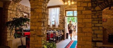 A stone-walled interior with a wooden ceiling, a red and blue patterned carpet, and a dining area with tables and chairs.