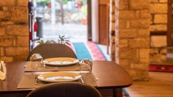 Un comedor con pared de piedra, una mesa de madera dispuesta para dos personas y vista a un espacio exterior con vegetación a través de un vano abierto.