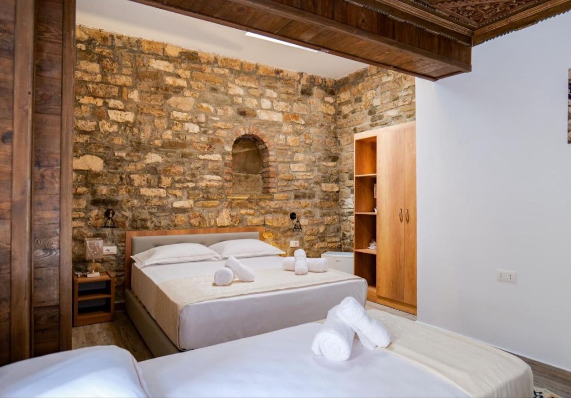 A room with two beds, a stone wall, wooden beams, a wooden wardrobe, and a small table with a lamp.