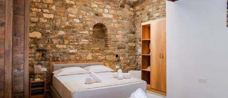 A room with two beds, a stone wall, wooden beams, a wooden wardrobe, and a small table with a lamp.