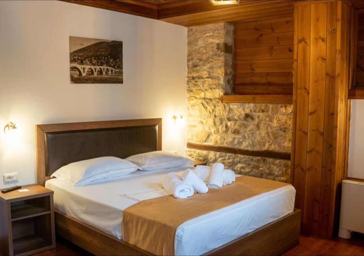 A hotel room with a stone wall, wooden ceiling, a bed with white linens, and a nightstand with a lamp.