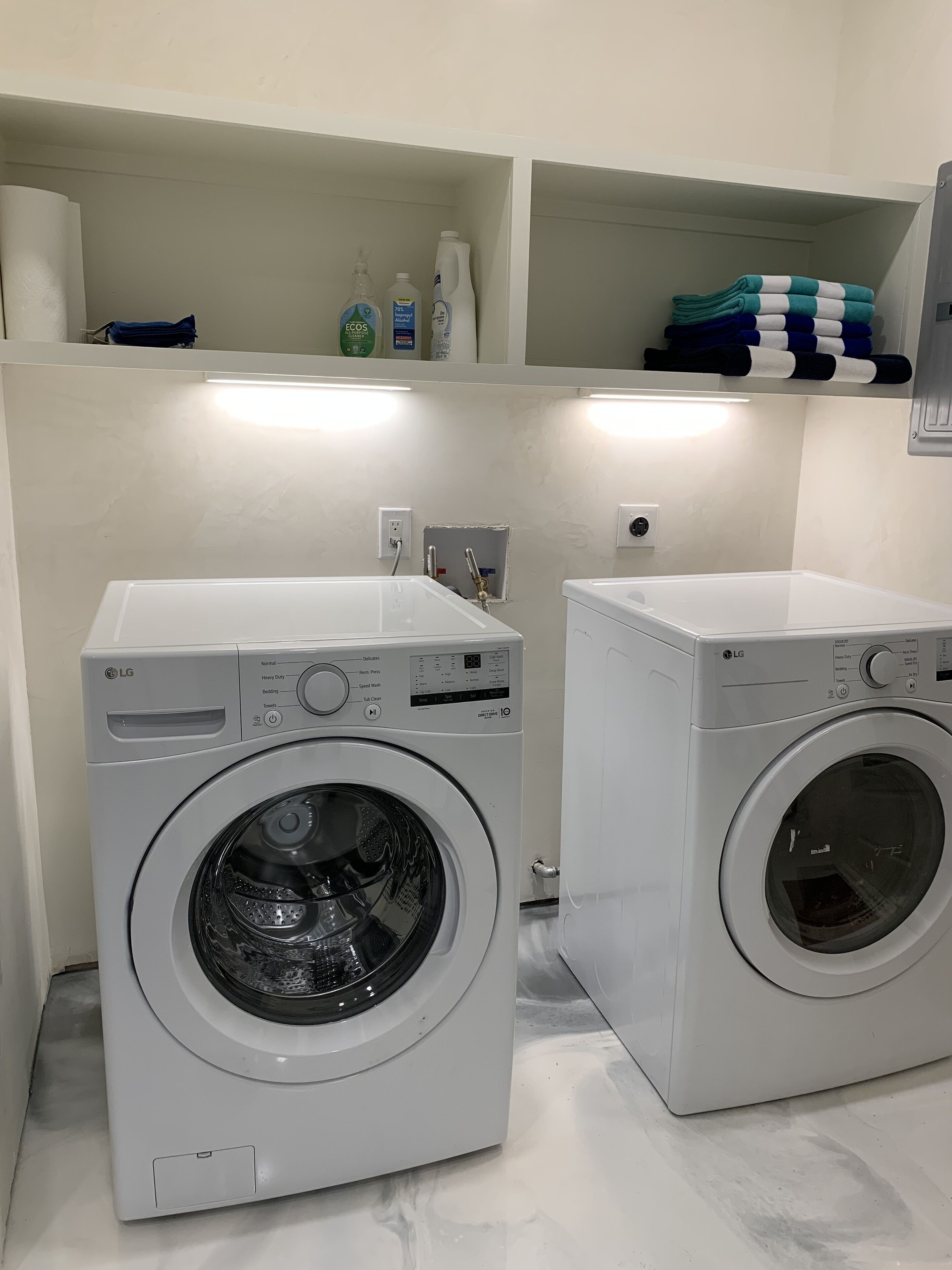 Dos lavadoras LG blancas de carga frontal en un cuarto de lavado con pared de azulejos blancos, un estante con toallas dobladas y algunas botellas de productos de limpieza.