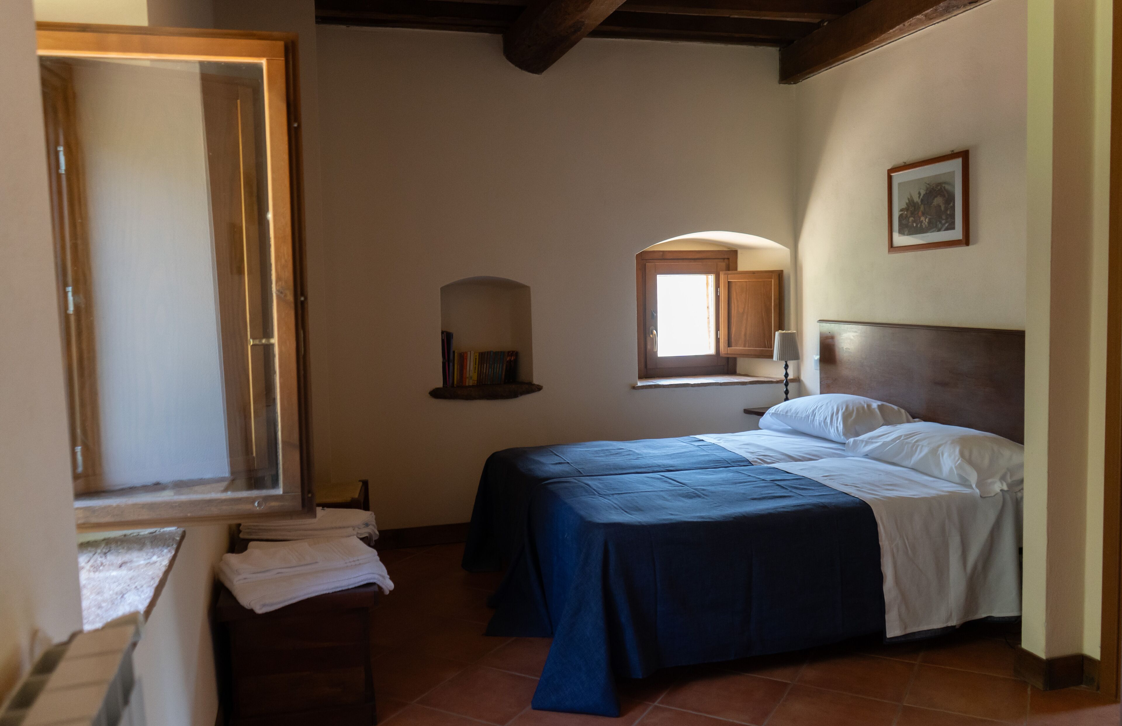 A bedroom with a bed, a window, a wooden headboard, a shelf with books, and a framed picture on the wall.