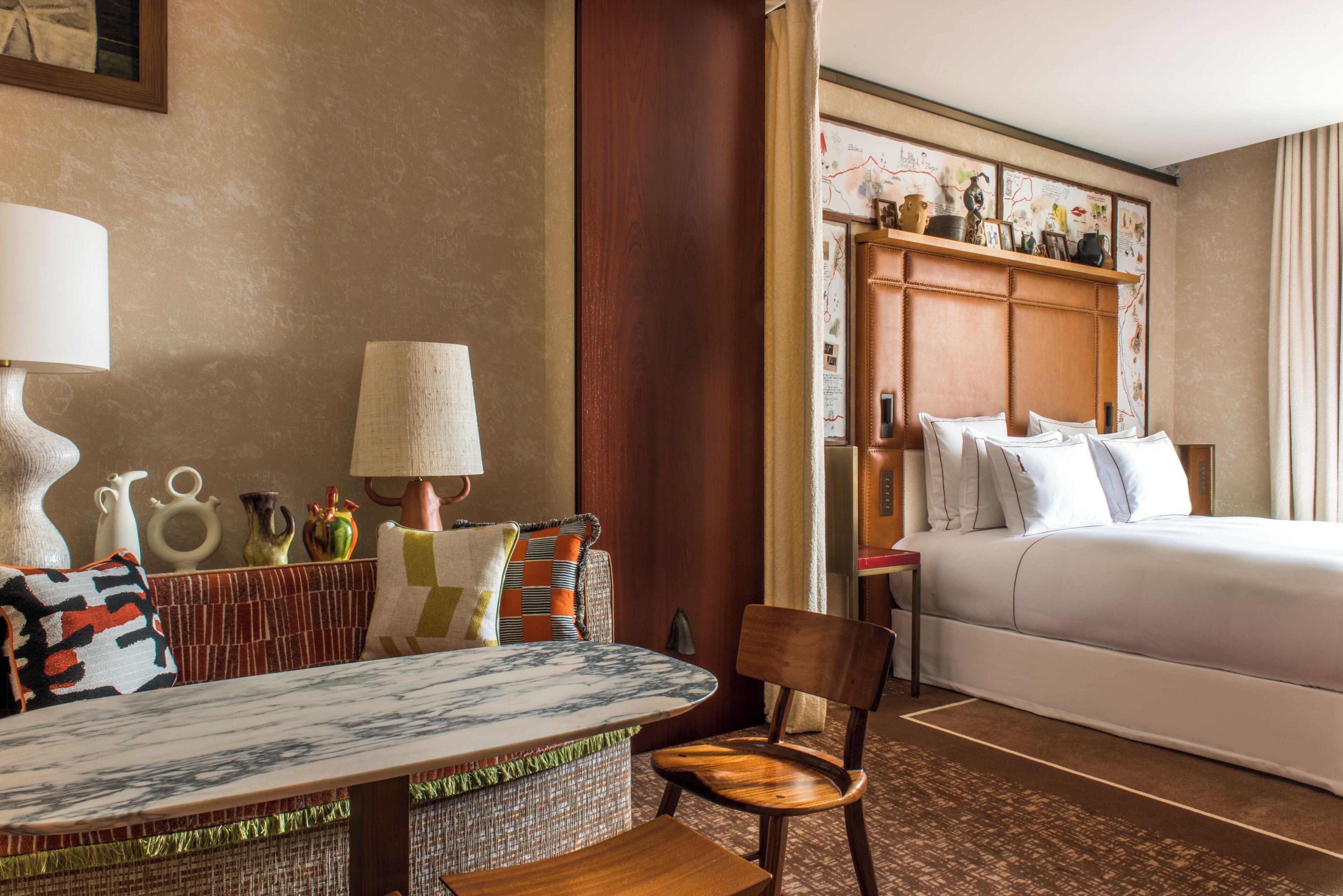 A hotel room with a bed, a wooden headboard, a dining table with chairs, a lamp, and various decorative items.