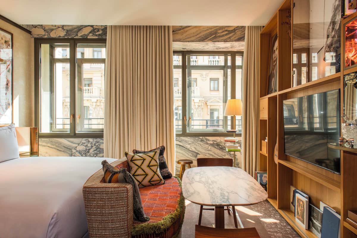 A hotel room with a large bed, a sofa with patterned cushions, a marble-topped table, a television, and a view of buildings through large windows.