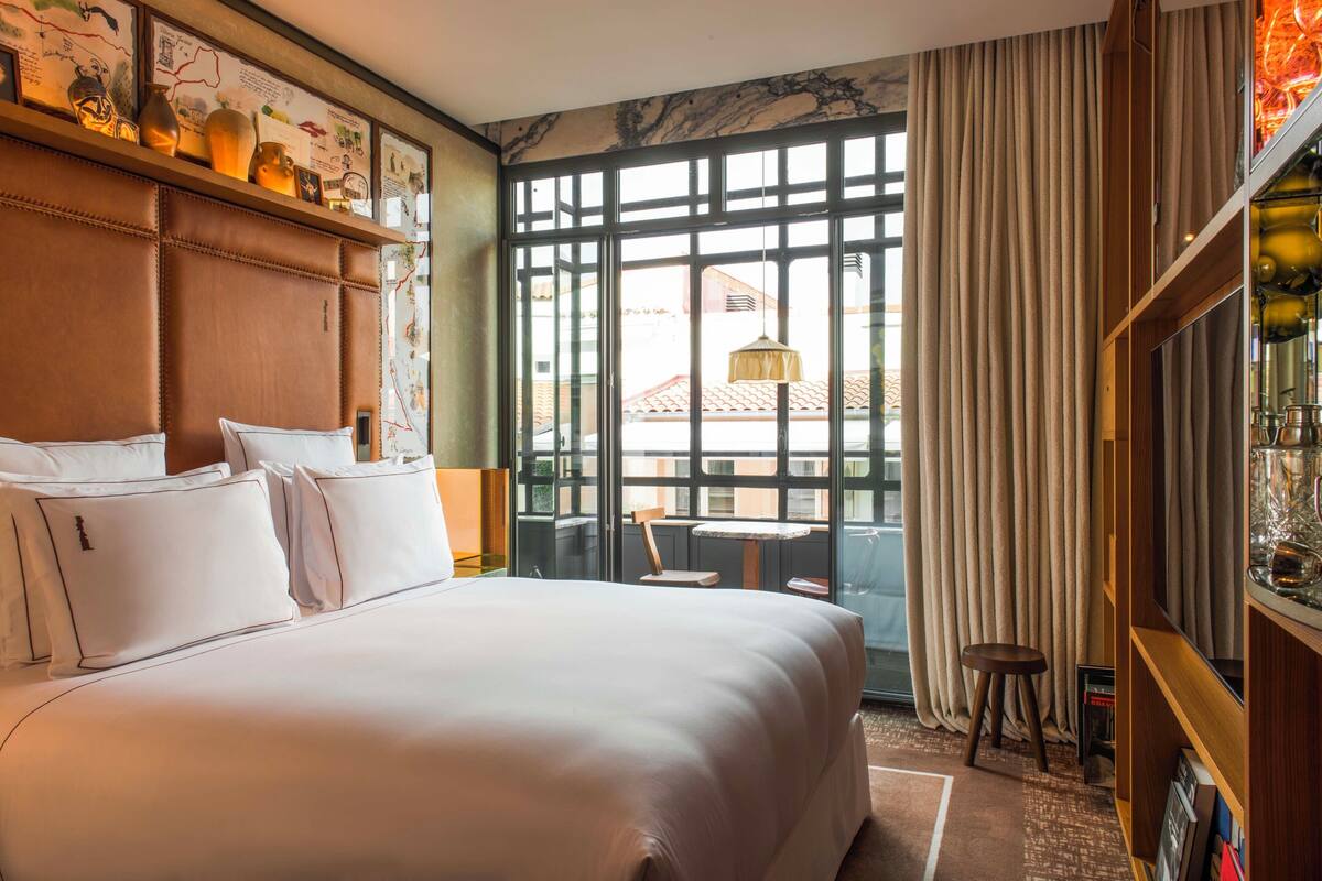 A hotel room with a large bed, a wooden headboard, a small table, and a view of the outside through large windows.