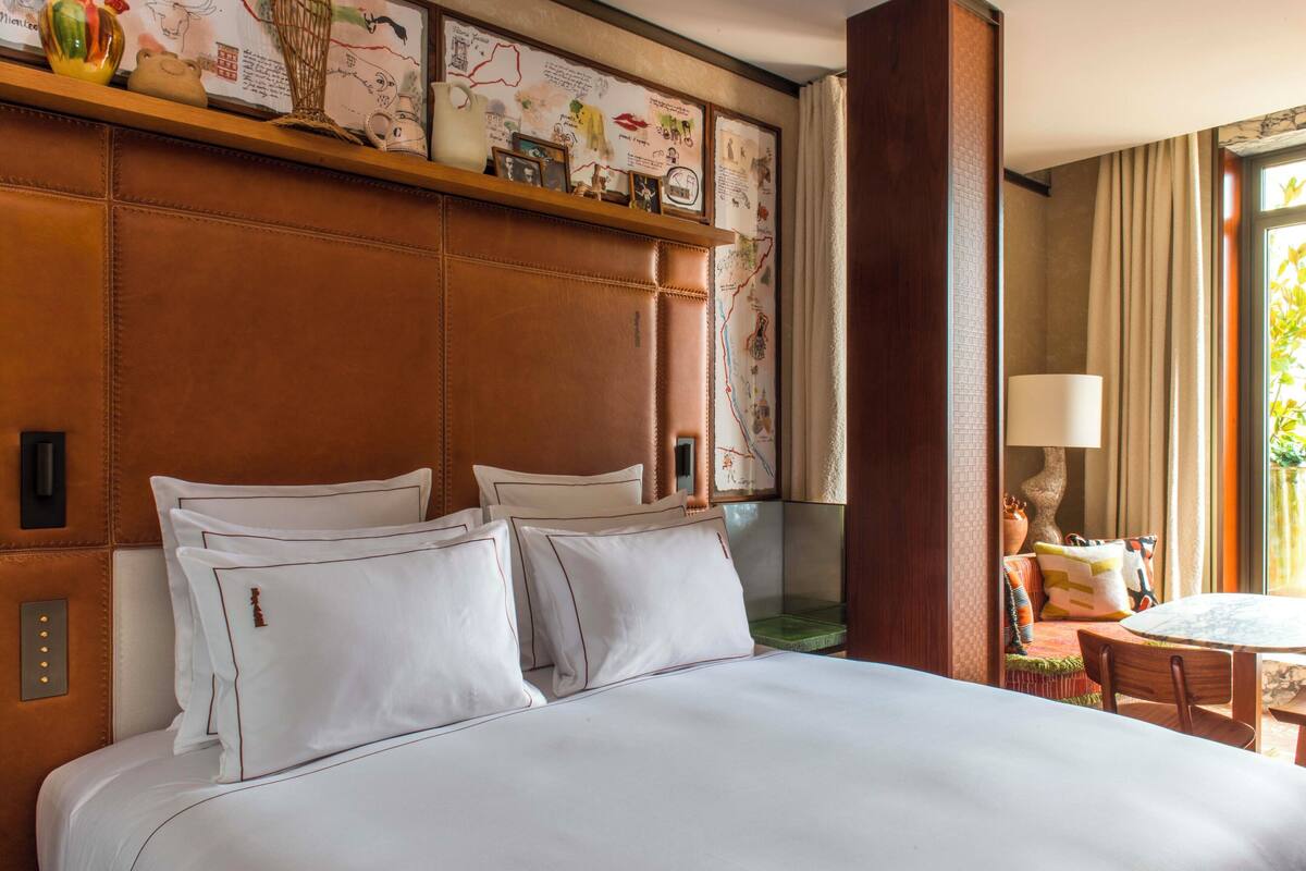 A hotel room with a large bed, a wooden headboard, a small table with a lamp, and a window with curtains.