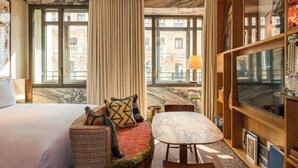 A hotel room with a large bed, a sofa with patterned cushions, a marble-topped table, a television, and a view of buildings through large windows. - Brach Madrid - Evok Collection (Madrid)